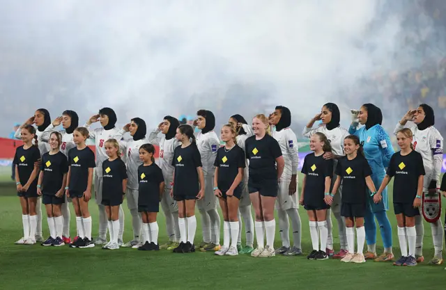 A row of women wearing white football kit salute while in front of them stand a row of girls wearing black t-shirts and shorts