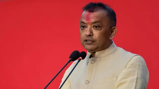 A man in a cream-coloured waistcoat and shirt speaks into a microphone in front of a red background