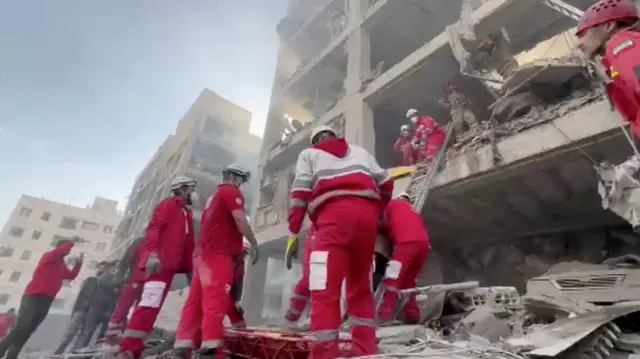 Lots of emergency workers stood at the bottom of a building among rubble. The building is very damaged with no walls