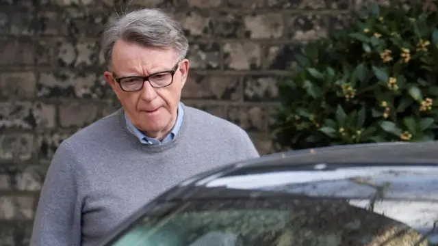 Peter Mandelson climbs into a car. He wears a grey jumper, blue shirt and black framed glasses