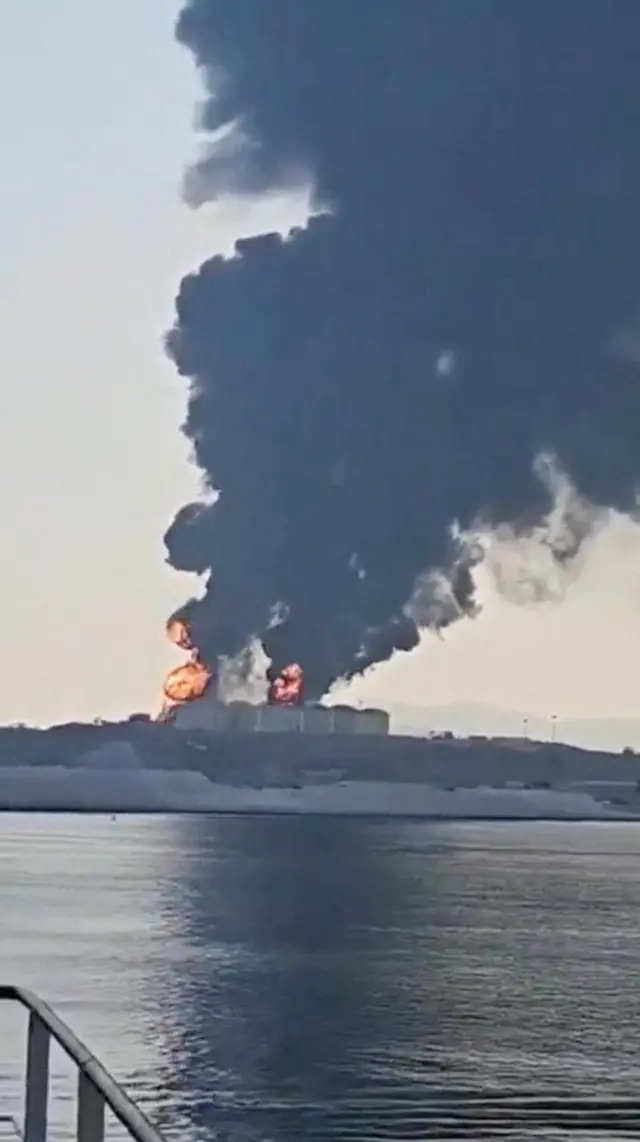 Black clouds of smoke rise from buring oil storage tanks by a body of water with fire and explosions also visible