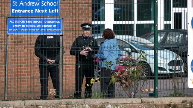 Two police officers talking to a woman wearing a blue jacket. They are standing on the other side of a green fence at the school.