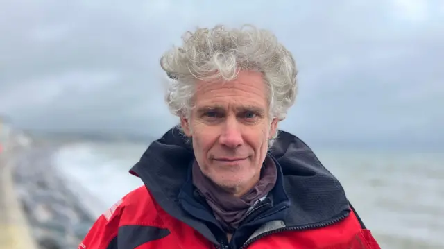 A man stands on the seafront at Torcross, Devon. There are waves crashing against the sea defences in the background. He is wearing a red waterproof jacket.