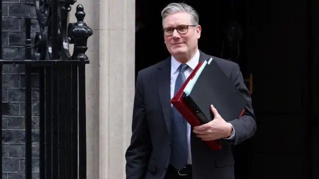 Keir Starmer walking out of 10 Downing Street holding a number of files. He is wearing a dark suit and grey tie