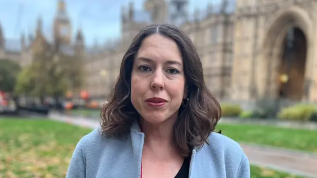 Alice MacDonald is standing outside the Palace of Westminster. She has brown hair, is pursing her lips, and is wearing a grey or sky blue coat.