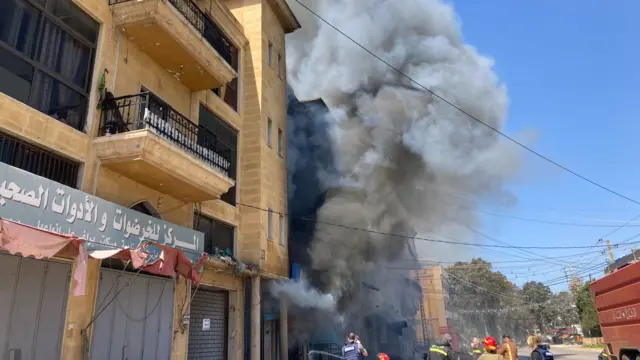 Smoke comes out of the side of a building while firefighters stand nearby