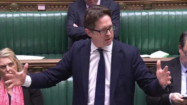 Alex Burghart with his arms and fingers stretched wide to either side, is speaking in the benches at the House of Commons for the Conservatives