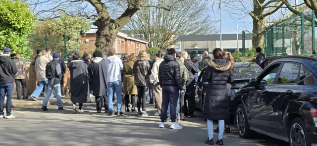 Crowds of people gathered outside Thorpe St Andrew School in Norwich. They all have their backs to the camera and there are cars parked on the side of the road.