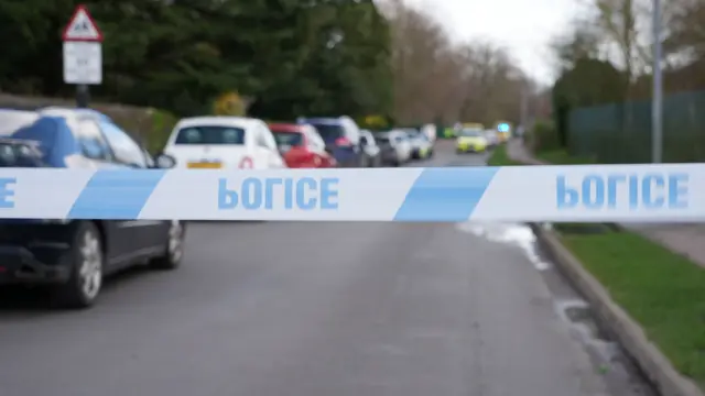 Blue and white police tape cordons off the road, which is lined with parked cars. They include police and an ambulance.