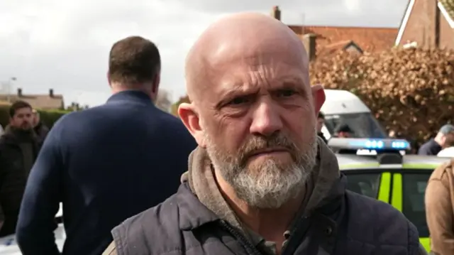 Adam, a man who is standing on Laundry Lane in Norwich. He is looking just off camera and is wearing a grey hoodie and coat.