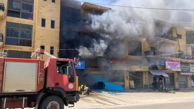 Thick black smoke comes from a building with a fire engine in the foreground. The building hit appears to have ships and a cafe on the ground floor