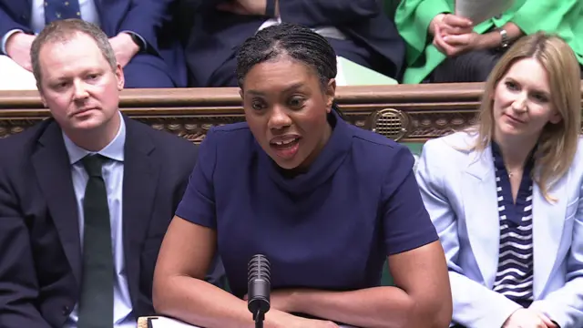 Kemi Badenoch wearing a blue t-shirt and speaking in the House of Commons