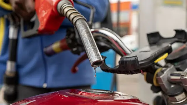 A pump attendant refuels a red motorcycle at a petrol station. A drop of petrol slides off the nozzle.
