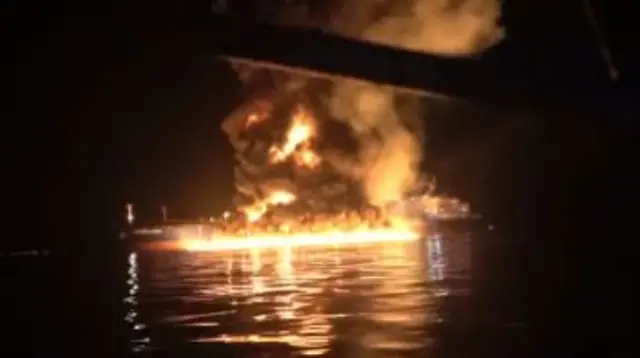 A ship at sea at night, engulfed by flames, with a large cloud of black smoke rising. Another ship is in close proximity