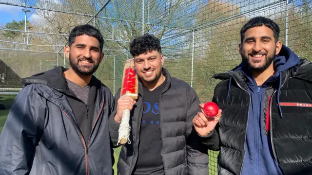 Abbas Sohail, Idrees Maskeen and Abdullah Sohail are standing from left to right smiling at the camera. They are all Asian men and all wearing dark blue or black jumpers and jackets with similar coloured coats. Idrees in the middle is holding a cricket bat over his right shoulder and Abdullah on the right is holding a red cricket ball in his right hand. Behind them are some cricket nets and trees.