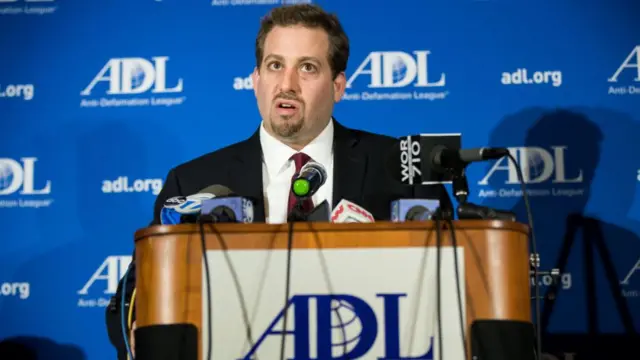 A man wearing a dark suit and maroon tie stands in front of a podium full of microphones. Signs behind him and on the podium read "ADL"