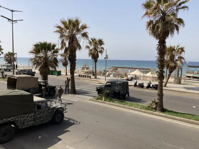 Four military vehicles on the coast of Beirut, Lebanon, which have camouflage pattern and palm trees growing either side of the roads and with the sea in the background