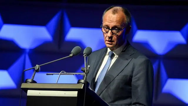 German Chancellor Friedrich Merz speaks at a news conference into a podium with two microphones and a geometric blue and dark blue pattern behind him