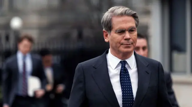 A man in a dark suit and spectacles walks outside in dim sunlight.