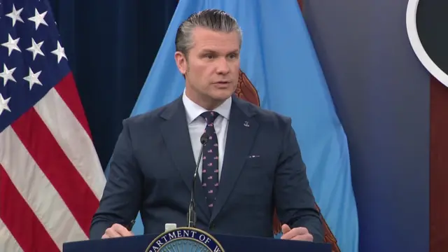 Ông Hegseth đang phát biểu giữa chừng phía sau bục giảng, phía sau là lá cờ Mỹ trên cột cờ.