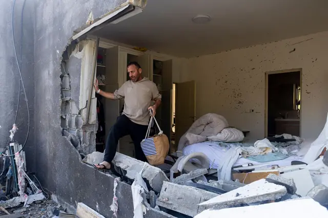 A man stands in a large hole in the side of a building, he is holding a small bag of belongings