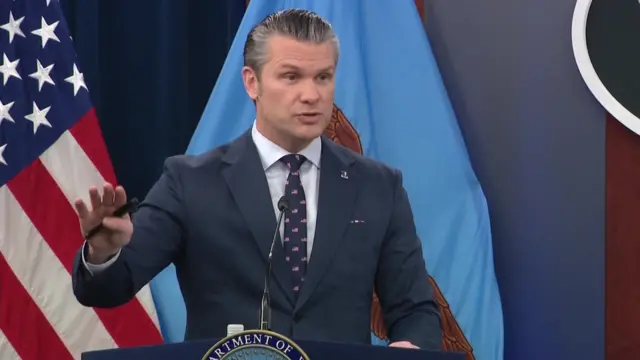 Pete Hegseth trong bộ vest màu xanh đậm, áo sơ mi trắng và cà vạt họa tiết đang phát biểu sau bục giảng.