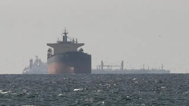 A ship sailing in the Straits of Hormuz with more infrastructure behind it slightly obscured in the foggy distance