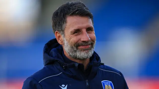 A smiling Colchester boss Danny Cowley wearing a blue training top with the club badge of an eagle on it