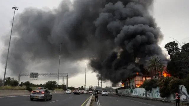 Thick plumes of smoke in the sky next to a building on fire