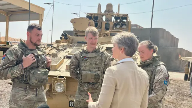 Foreign Secretary Yvette Cooper speaking to three soldiers in camouflage gear in front of a military vehicle on a sunny, dusty day in Saudi Arabia