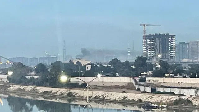 Smoke rises in the distance of a city landscape with a high rise building next to a crane, and water at the bottom of the frame.