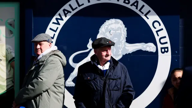 Millwall fans stand in front of a giant club crest at The Den