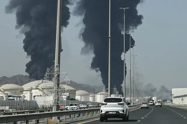 Two plumes of black smoke rise among white siloes on the side of a wide highway where white cars are also driving
