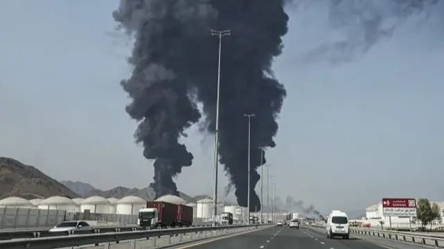 Two huge pillars of black smoke rise in the distance near a motorway in front of some mountains. A number of white tanks can be seen in front of the smoke