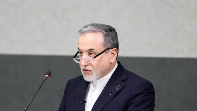 Araghchi speaking into a small microphone that's on a stand in front of him. Only his shoulders up are visible. He wears a suit and glasses. The background is two tones of grey.