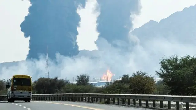 Two columns of thick, black smoke are seen from the side of the road. Bright, tall flames can be seen in front of one of the plumes of smoke. Several foothills can be seen in the background