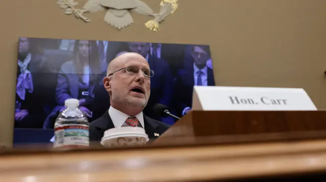 Federal Communications Commission (FCC) Chairman Brendan Carr testifies before a hearing with a TV screen behind him and a water bottle, coffee and name card in front.