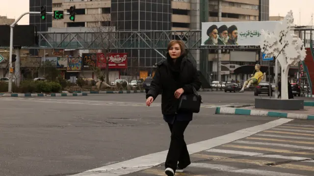 A woman walks across the street, with a banner of all three leaders of Iran, late Ruhollah Khomeini, late Ayatollah Ali Khamenei, and Mojtaba Khamenei in the backgroun