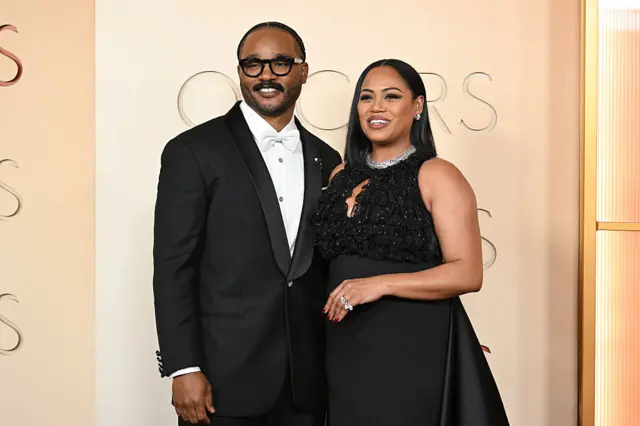 Ryan and Zinzi Coogler pose for a photo in a black tuxedo with a white bowtie and a black dress, respectively