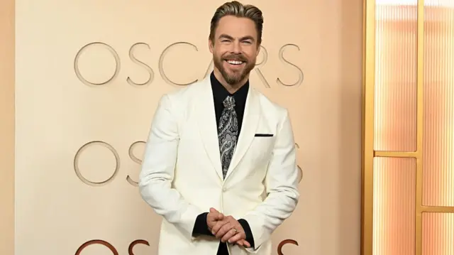 Derek Hough, in a white suit and black shirt, smiling for a photo