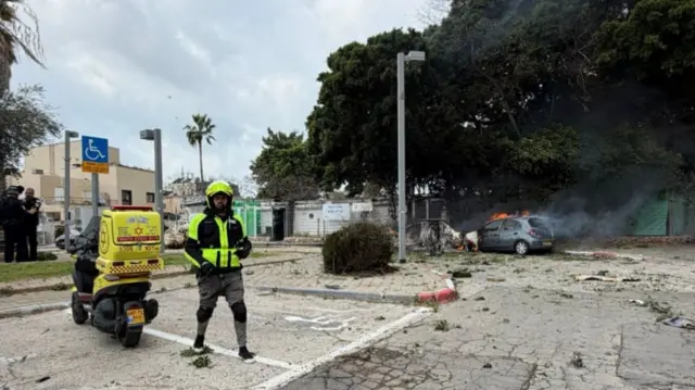 An emergency services worker stands next to a motorbike in front of a burning car