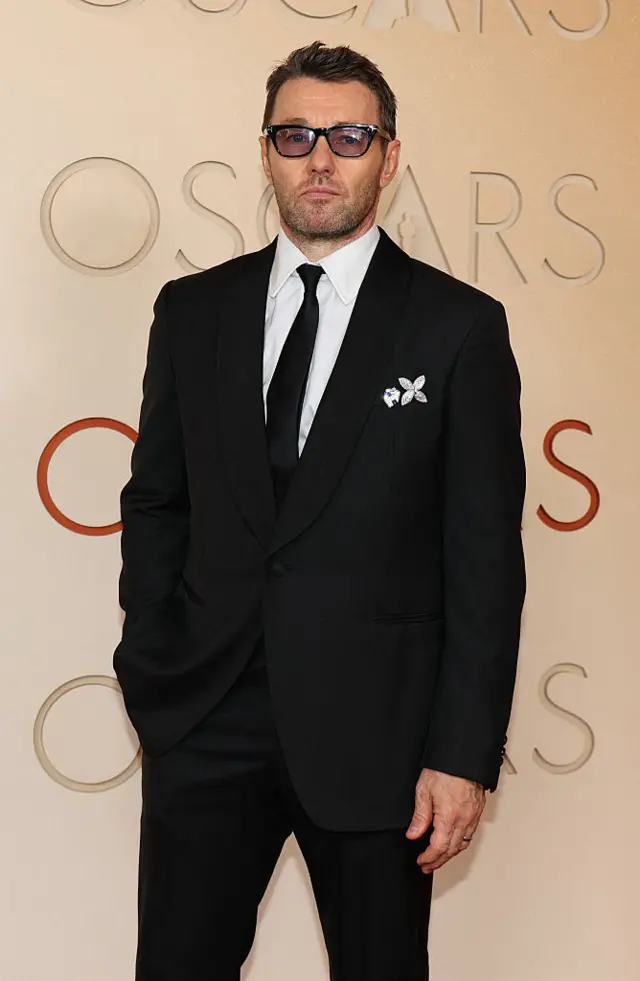 Joel Edgerton in a black suit, white shirt and white tie, with sunglasses