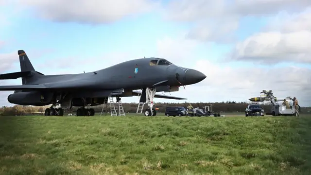 A USAF B1 B bomber stands on the grass at an RAF base. A number of workers stand near a large missile nearby