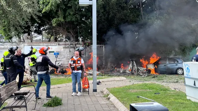 Emergency services respond at scene of fire following latest Iranian strikes on Israel. A car is seem burning and smoke is rising.