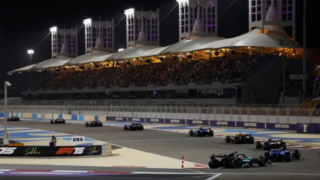 Formula One cars race along a track in front of a grandstand at night.