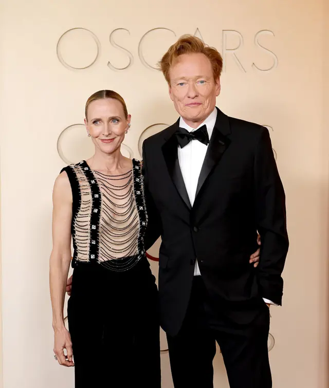 Conan O'Brien and his wife Liza pose for a photo. Conan wears a tuxedo and his wife wears a black and beige dress adorned with chains