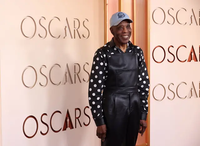 Buddy Guy in black leather dungarees, a polka dot shirt and blue baseball cap