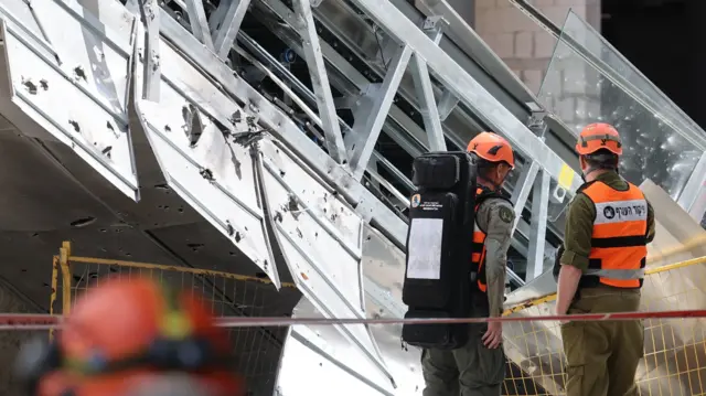 Two men in high vis stand near a cordoned off building that's damaged