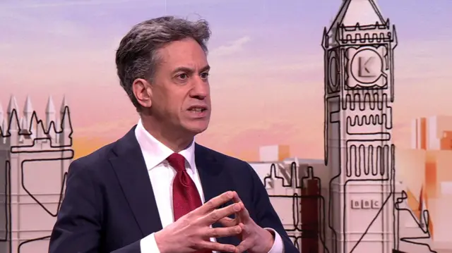 Ed Miliband in dark blue suit, white shirt and red tie sitting down in a TV studio, his hands in front of him