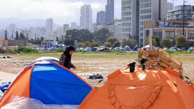 Two tents can be seen in front of a view of the city Beirut with high rise buildings. A woman walks behind one of them